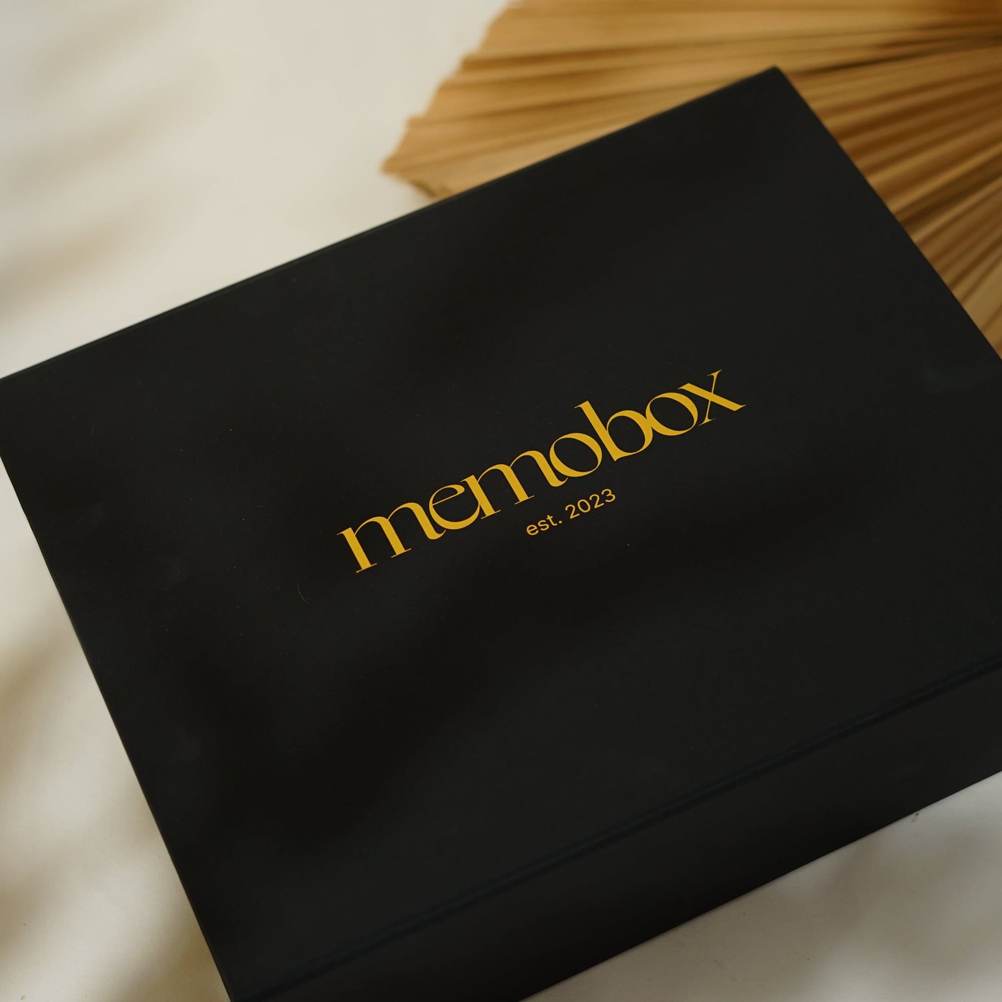 The black Memobox lid, gold-stamped, on a pale ground beside a dried palm fan
